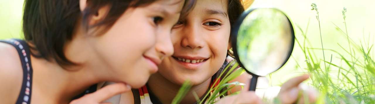 Curious kids using a magnifying glass to look at grass in a garden.