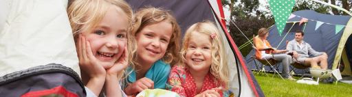 Three girls sitting in a tent camping while their parents sit nearby.