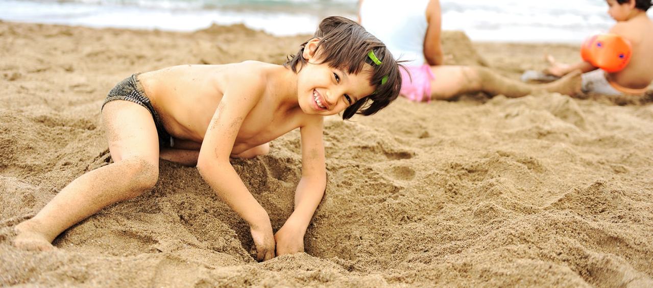 Happy boy digs a hole in the sand on a beach while other children play nearby.