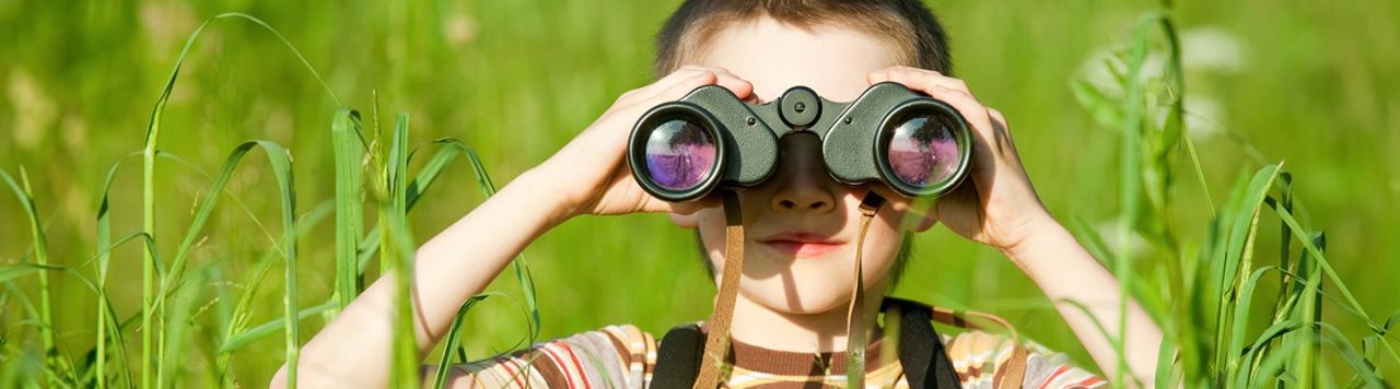 Boy looking through binoculars in a meadow to find scavenger hunt clues.