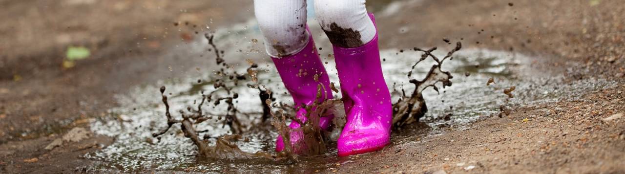 Child splashing in mud puddles with pink wellies and mud stains on her leggings.
