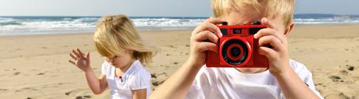 A boy holds up a camera while his sister plays on a beach.